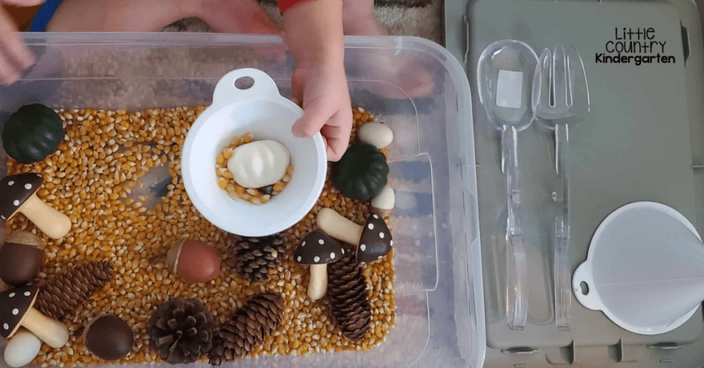 Fall sensory bin with popcorn kernels, a funnel with a pumpkin in it, and a lid with the tools organized on it.